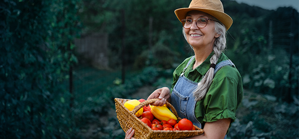 Femme souriante tient un panier de légumes dans un jardin verdoyant.